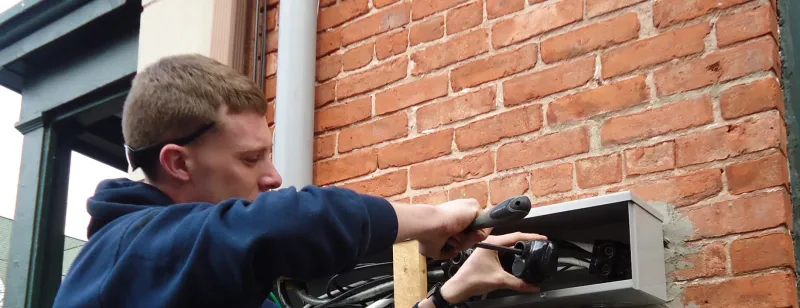 An electrician connects wiring at a meter box on a brick wall.